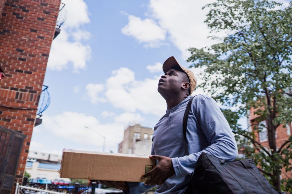 Man delivering a package in a city neighborhood, looking up at the building.